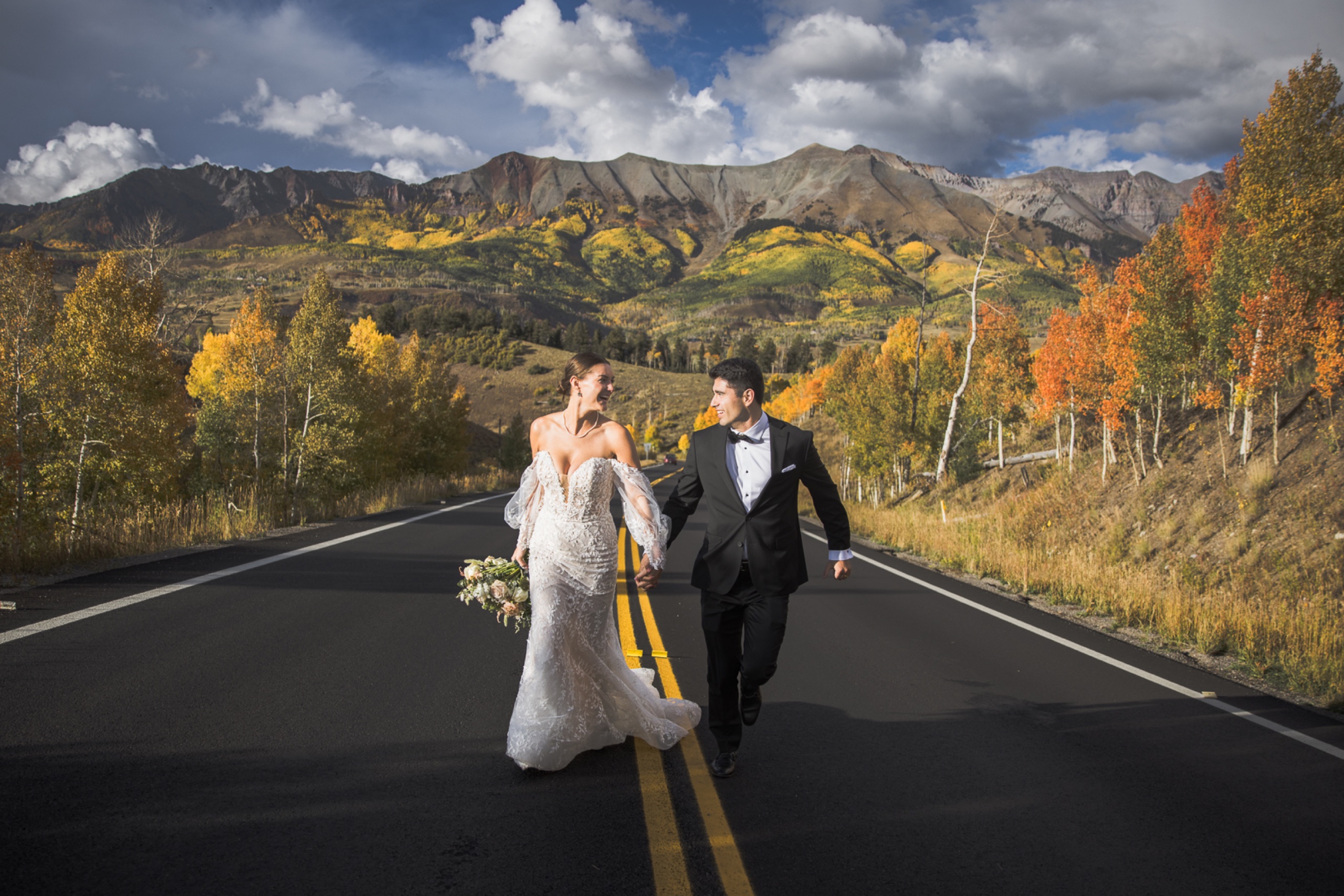 Bride and groom walking hand in hand down a mountain highway surrounded by vibrant fall foliage in Telluride, Colorado