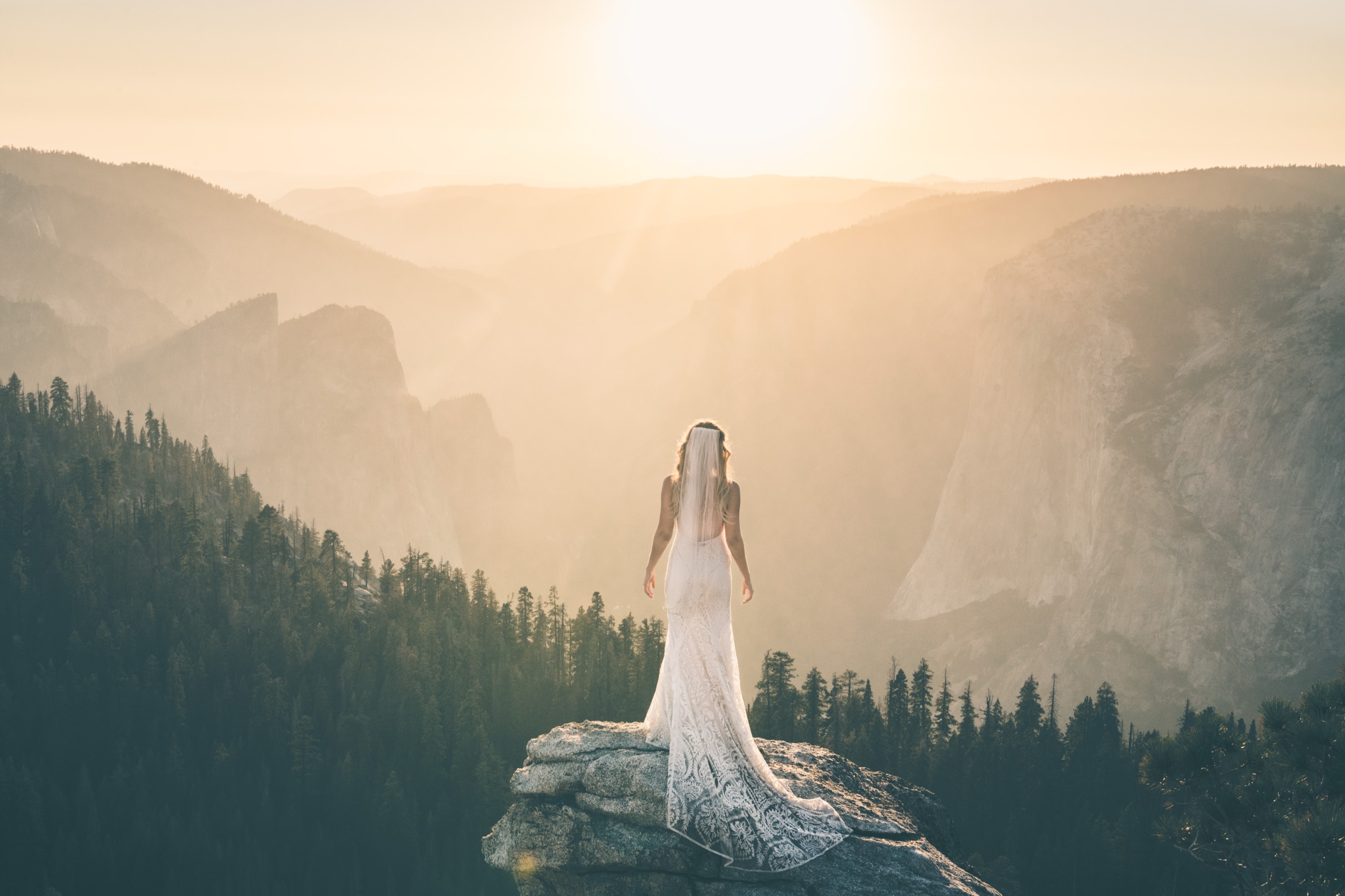 Bride in a lace gown standing alone on a granite cliff overlooking Yosemite Valley at golden hour sunset