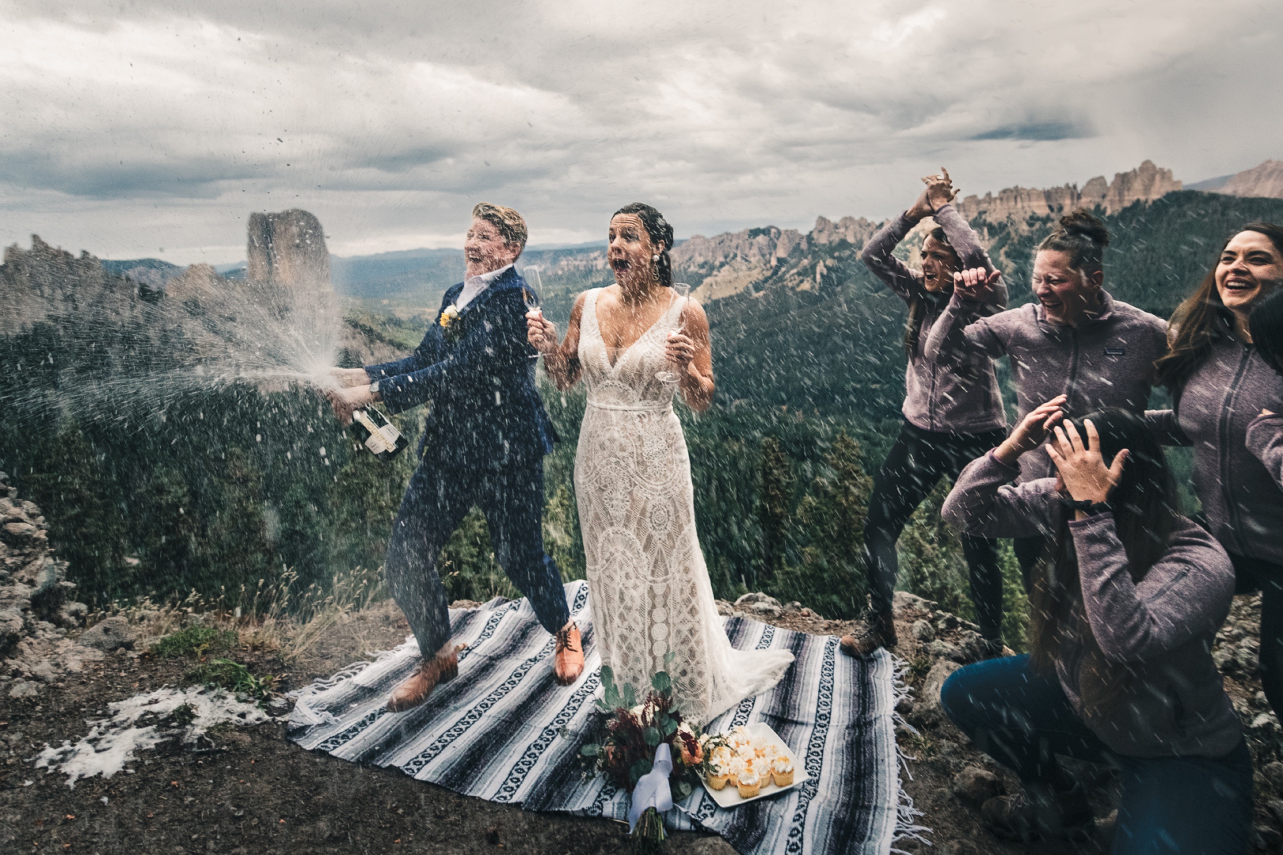 Wedding couple celebrating with a champagne spray on a rugged mountain summit surrounded by their guests