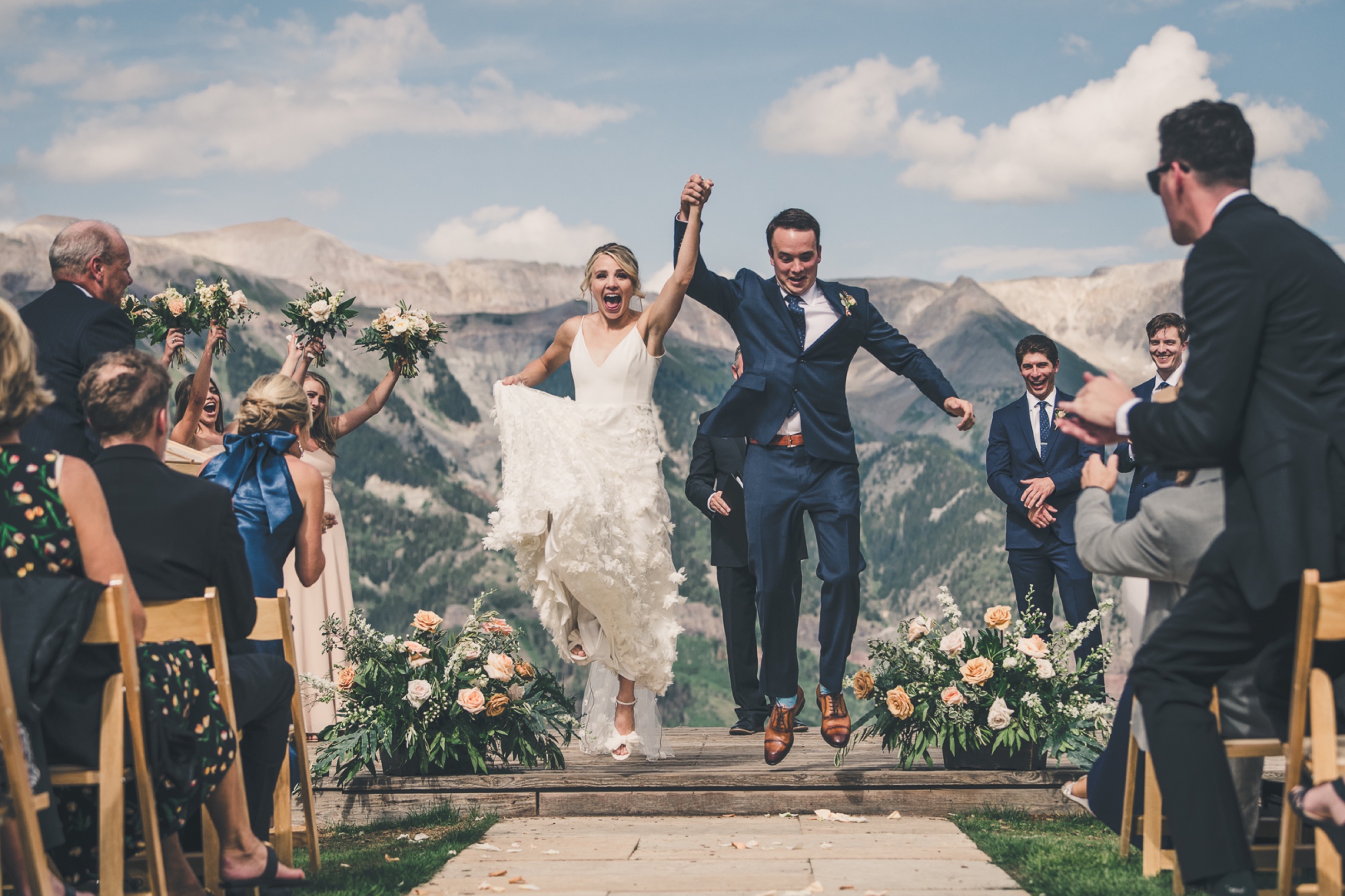 Ecstatic bride and groom jumping together as they walk the aisle with Telluride mountain peaks behind them