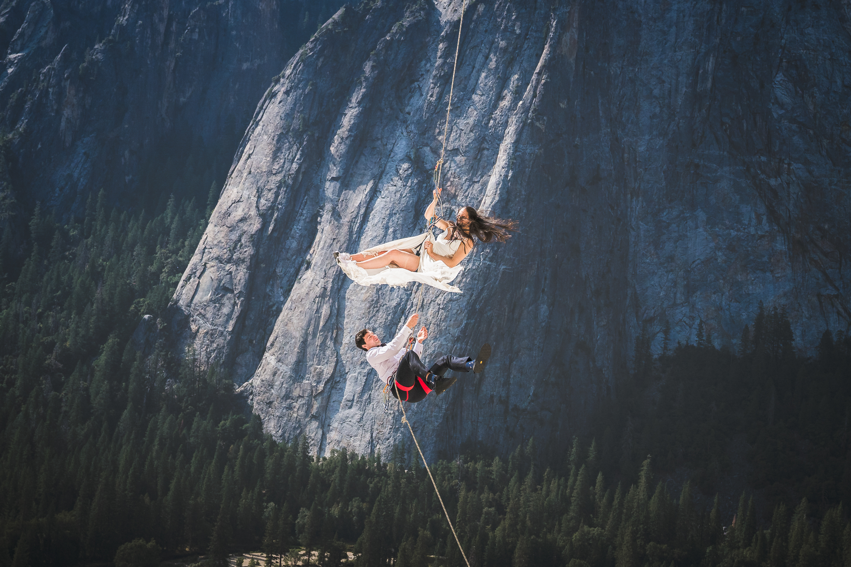 Bride and groom rappelling down the sheer granite face of El Capitan in Yosemite in wedding attire