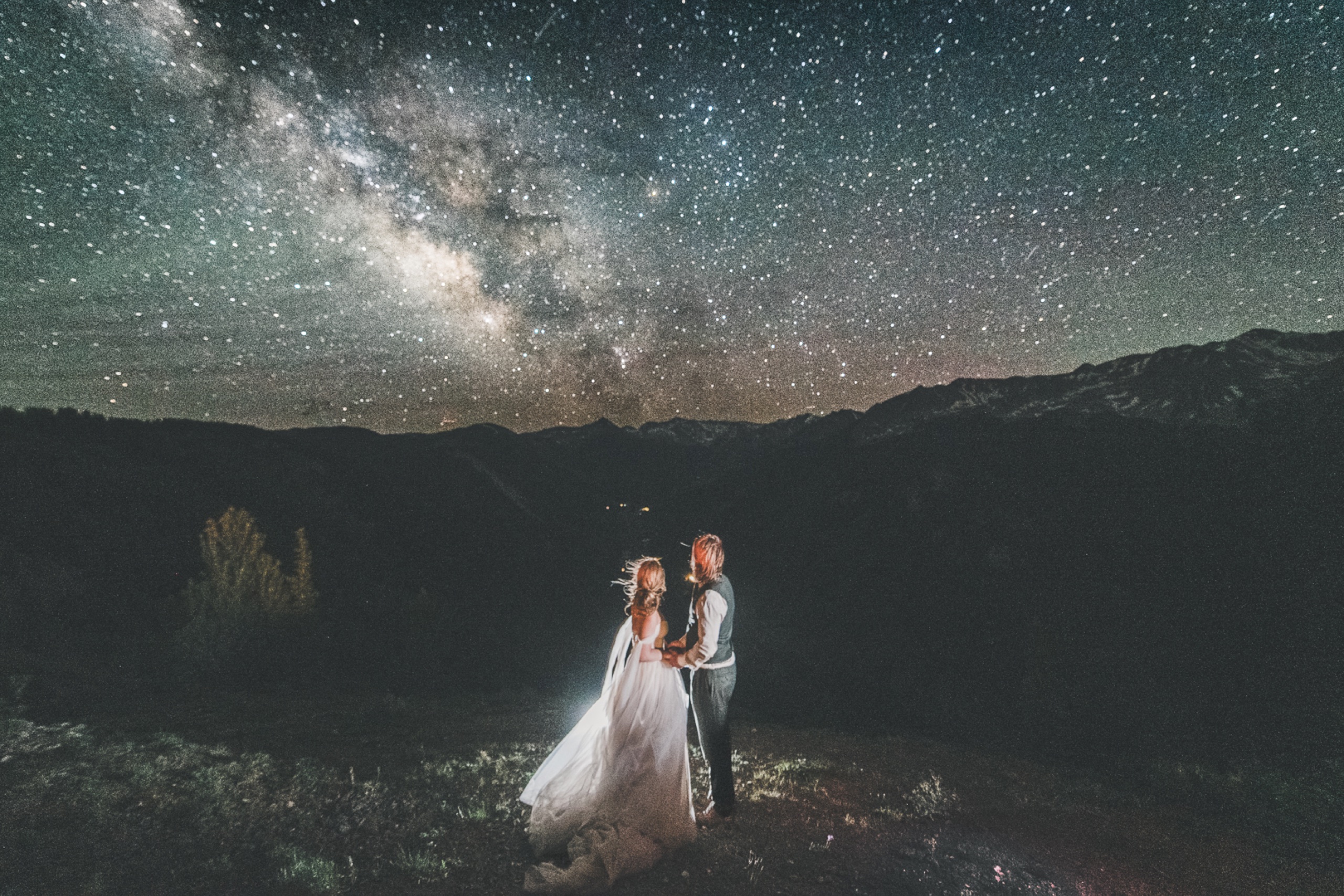 Wedding couple standing beneath the Milky Way galaxy on a mountaintop, lit by a single warm light source