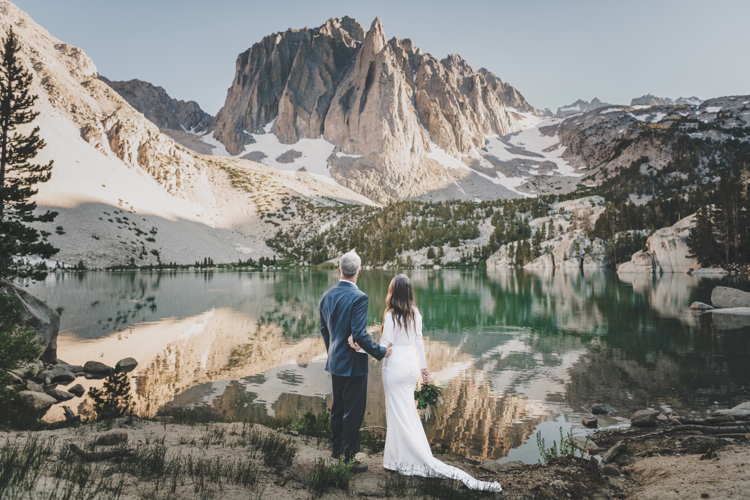 Couple standing hand in hand before a mirror-still alpine lake reflecting jagged Sierra Nevada peaks in the Eastern Sierra